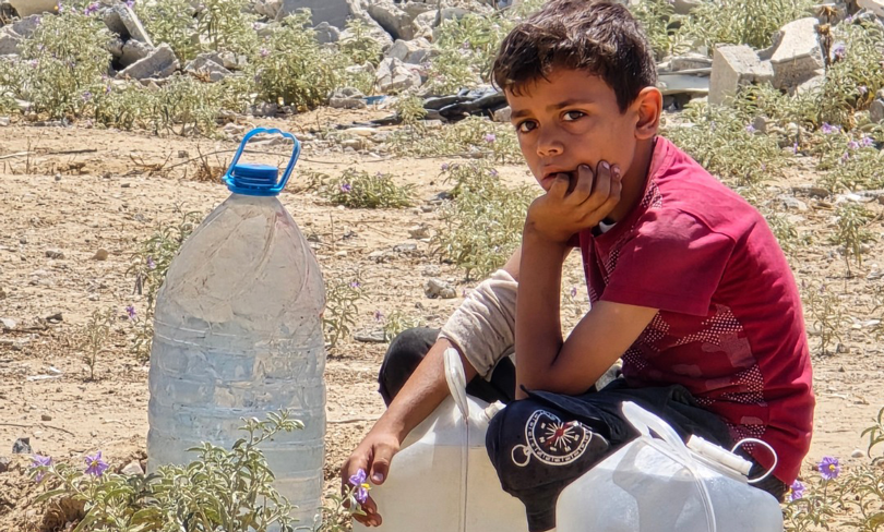 A young boy in Gaza sits next to empty water containers